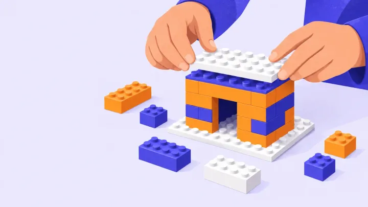 Hands place a white roof on a small orange and indigo brick house beside loose toy bricks